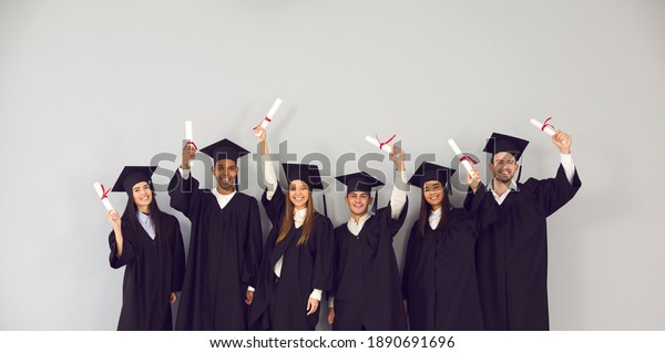 Study abroad website banner. Group of happy\
smiling diverse academy graduates holding up diplomas.\
International university students in traditional black academic\
gowns and caps celebrating\
graduation