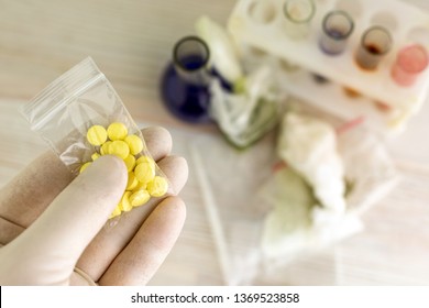 Study Of Synthetic Drugs: A Scientist Holds A Sample Of A New Drug Above The Table With Other Samples, Short Focus