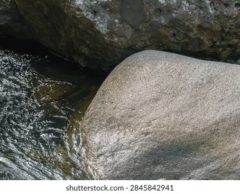 A study of texture and scale, featuring two massive boulders side-by-side at the edge of a clear river. One rock has prominent green moss, the other is a rougher grey. A powerful image of natural coexistence and raw material. Useful for geology, environmental studies, and natural history.