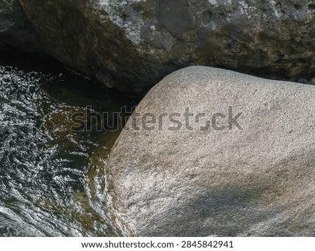 A study of texture and scale, featuring two massive boulders side-by-side at the edge of a clear river. One rock has prominent green moss, the other is a rougher grey. A powerful image of natural coexistence and raw material. Useful for geology, environmental studies, and natural history.