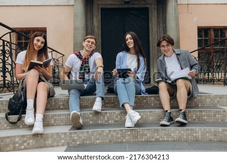 Study together is fun, teamwork, team building concept. Four happy students are sitting near college building and holding books, devices, wearing casual smart, smiling, on a nice summer