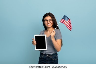Studying abroad online. Happy Indian teen female student showing tablet pc with empty screen, holding American flag, receiving education or working in United States on web, blue background. Mockup