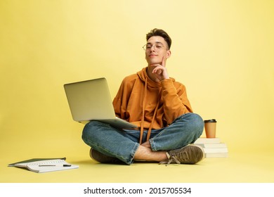 勉強や宿題。黄色いスタジオ背景にグラスをかけた学生が床に座り、ノートパソコンを持つ。教育、勉強、生活のコンセプト。