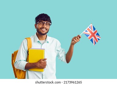 Studying In England. Portrait Of Happy Indian Male University Student With UK Flag On Light Blue Background. Ethnic Smart Guy With Backpack And Notebooks Waving England Flag Looking At Camera.