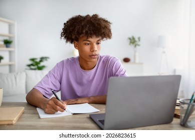 Studying From Home Concept. Serious Black Teen Guy Taking Notes While Participating In Online Lesson On Laptop. Cool African American Youth Making Homework, Speaking To His Tutor On Web