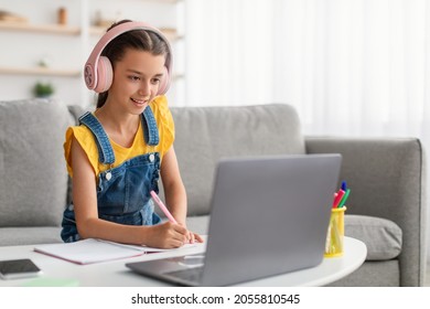 Studying At Home. Smiling pupil in pink headset sitting on couch at table using pc, writing essay or test exam, taking notes in textbook listening to teacher looking at screen during online lesson - Shutterstock ID 2055810545