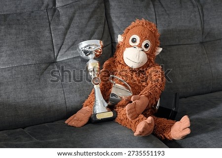 A stuffed monkey is sitting on a couch holding two cups and two trophies. The monkey is smiling and he is proud of its accomplishments
