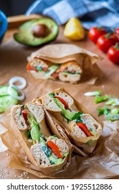 Stuffed Toasted Bagel Sandwich Cut Into Half With Avocado, Mayo, Cucumber, Lettuce And Tomatoes. Salmon Bagel In The Background, Wrapped With Paper. Gluten Free Healthy Breakfast, Vibrant Fun Colors 