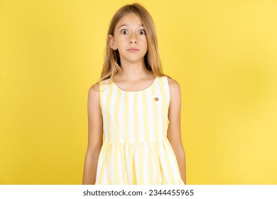 Stunned Caucasian kid girl wearing yellow dress over yellow background stares reacts on shocking news. Astonished preteen girl holds breath