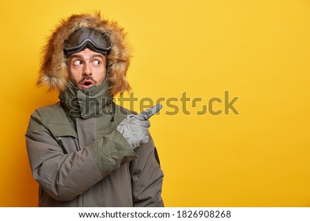 Stunned Caucasian man in winter clothes points away on blank space with surprised expression goes skiing during cold day wears jacket and gloves enjoys chilly weather isolated on yellow background