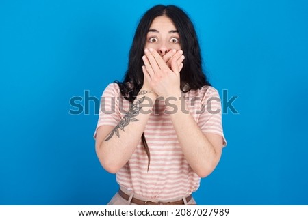 Stunned Caucasian woman wearing striped T-shirt over blue background covers both hands on mouth, afraids of something astonishing