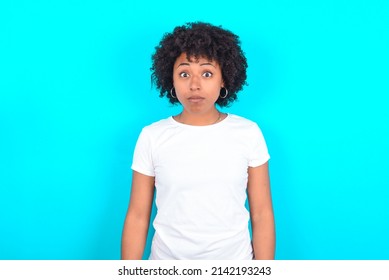 Stunned young woman with afro hairstyle wearing white T-shirt against blue wall stares reacts on shocking news. Astonished girl holds breath