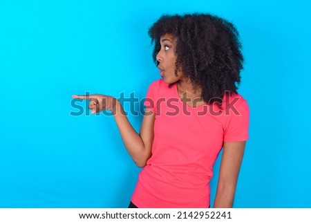 Stunned Young woman with afro hairstyle wearing pink t-shirt over blue background with greatly surprised expression points away on copy space, indicates something