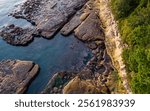 A stunning aerial photograph capturing the rugged coastline of Vancouver Island, British Columbia. The image shows detailed rock formations surrounded by clear blue ocean waters and lush green foliage