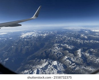 A stunning aerial view of the Alps, showcasing snow-capped peaks, rugged terrain, and a clear blue sky, captured from an airplane window. - Powered by Shutterstock