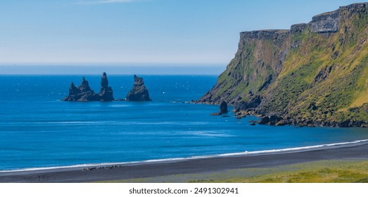 Stunning aerial view of a black sand beach in Iceland, bordered by rocky cliffs and distant sea stacks. The vibrant blue water and clear sky enhance the dramatic landscape. - Powered by Shutterstock