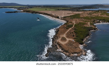 A stunning aerial view of a coastal landscape featuring a serene bay, vast green fields, and distant mountains under a clear blue sky. The image captures a peaceful seaside area with a few ships in th - Powered by Shutterstock