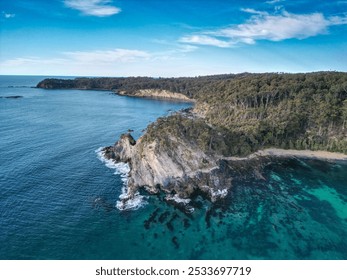 A stunning aerial view of a coastal landscape with cliffs, clear blue water, and lush green forest under a bright blue sky. - Powered by Shutterstock