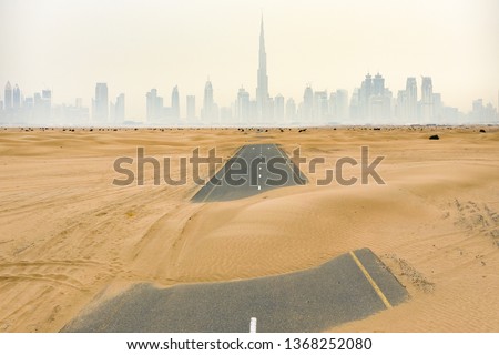 Stunning aerial view of a deserted road covered by sand dunes in the middle of the Dubai desert. Beautiful Dubai skyline surrounded by fog in the background. Dubai, United Arab Emirates.