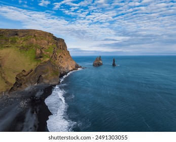 Stunning aerial view of Iceland's rugged coastline featuring a black sand beach, rocky cliff, and distant sea stacks under a bright blue sky with white clouds. - Powered by Shutterstock