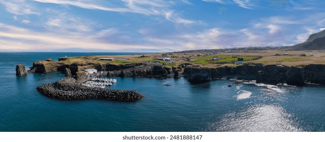 A stunning aerial view of a small harbor in Iceland, surrounded by rugged cliffs and green vegetation. The deep blue water contrasts with the bright blue sky and white clouds. - Powered by Shutterstock