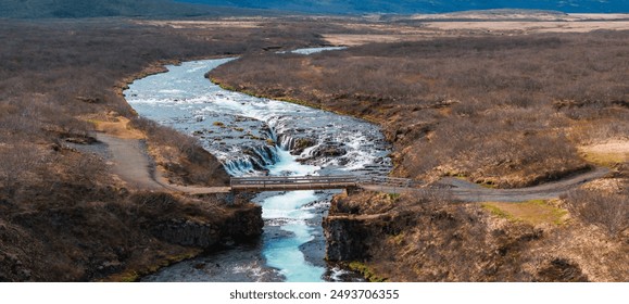 A stunning aerial view of a turquoise river flowing through a rugged landscape in Iceland, featuring a wooden bridge over a small waterfall and brown grass on either side. - Powered by Shutterstock
