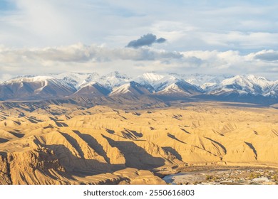 Stunning aerial view of a vast desert landscape with golden ridges and valleys, bordered by majestic snow-capped mountains under a dramatic sky. A breathtaking natural contrast of warm and cool tones. - Powered by Shutterstock