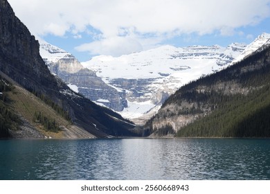 A stunning alpine lake surrounded by towering snow-capped mountains and lush forested slopes. The turquoise water reflects the dramatic cliffs under a bright blue sky. Lake Louise - Powered by Shutterstock