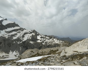 Stunning alpine landscape featuring dramatic snow-capped mountain peaks with exposed dark rock formations and patches of lingering snow. The composition showcases rugged terrain with large boulders in the foreground, sparse alpine vegetation, and rolling mountain ranges receding into the misty distance. Overcast sky with soft cloud cover creates moody atmospheric lighting typical of high-altitude European mountain environments. This image captures the raw beauty of the Alps during late spring or early summer transition season. Perfect for travel, nature, outdoor adventure, environmental, and geographical editorial and commercial uses.