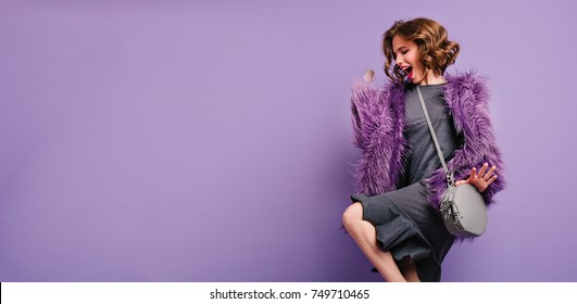 Stunning barefooted woman in trendy fur coat dancing and laughing on photoshoot. Indoor photo of lovely girl with short hairstyle fooling around on purple background.