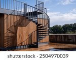 A stunning and beautifully designed spiral staircase gracefully ascends to an elevated area, which is enhanced by a warm wooden wall and a peaceful, inviting outdoor setting