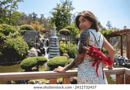 Stunning beauty and japanese style: brunette girl with tattoos, asian dress, and red paper umbrella, delighting in a sunny day with blue sky in a japanese garden