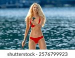 A stunning blonde bikini model glides across a serene lake on a paddleboard, basking in the warmth of the summer sun. The sparkling water and vibrant scenery add to the idyllic, sun-soaked vibe.