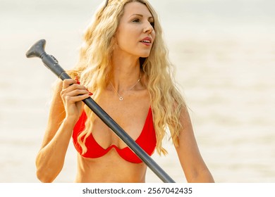 A stunning blonde bikini model glides across a serene lake on a paddleboard, basking in the warmth of the summer sun. The sparkling water and vibrant scenery add to the idyllic, sun-soaked vibe. - Powered by Shutterstock