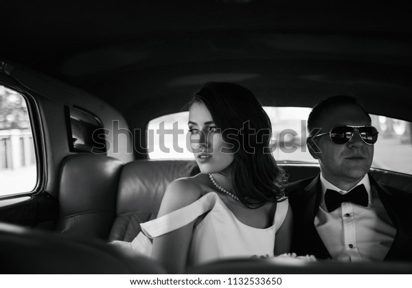 Stunning bride and groom in black sunglasses sit\
inside a retro car