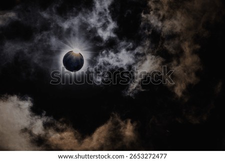 A stunning capture of a total solar eclipse as the moon passes directly in front of the sun, revealing a radiant corona against a dramatic cloud-filled sky. The surrounding clouds add a mystical, mood