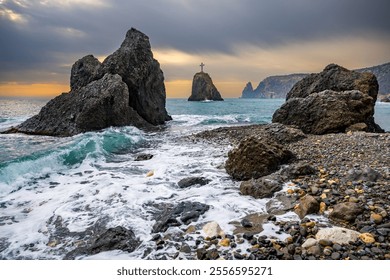 A stunning coastal landscape featuring rugged rocks and a pebble-covered shoreline. The ocean waves crash against the shore, creating white foam, while a dramatic sky, tinged with warm hues of sunrise - Powered by Shutterstock