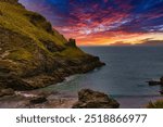 A stunning coastal landscape at sunset, featuring rocky cliffs and a vibrant sky filled with orange, pink, and purple hues. At Tintagel, Cornwall, UK.