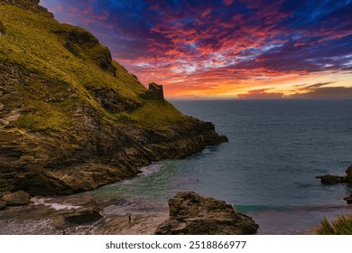 A stunning coastal landscape at sunset, featuring rocky cliffs and a vibrant sky filled with orange, pink, and purple hues. At Tintagel, Cornwall, UK. - Powered by Shutterstock