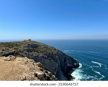 A stunning coastal view of a rocky cliff overlooking the vast, deep blue ocean. A small building sits perched on the hilltop, while the sea gently crashes against the rugged shoreline under the sky. - Powered by Shutterstock