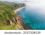 The stunning coastline displays rugged cliffs meeting a clear blue sea, with a tranquil sandy beach nestled amid lush greenery, enveloped in morning mist.Vladivostok russian far East