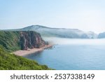 The stunning coastline displays rugged cliffs meeting a clear blue sea, with a tranquil sandy beach nestled amid lush greenery, enveloped in morning mist.Vladivostok russian far East