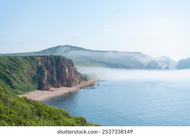 The stunning coastline displays rugged cliffs meeting a clear blue sea, with a tranquil sandy beach nestled amid lush greenery, enveloped in morning mist.Vladivostok russian far East - Powered by Shutterstock