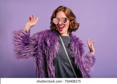 Stunning Cute Girl With Curly Brown Hair Posing With Pleasure On Purple Background. Indoor Photo Of Dancing Young Lady In Trendy Fur Coat Laughing To Camera.