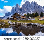 Stunning Dolomites, a quaint wooden cabin stands beside a tranquil pond, reflecting the imposing mountain ranges in the background.Geisleralm Dolomites Val Di Funes in Italy Adolf Munkel Trail Autumn