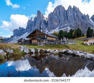 Stunning Dolomites, a quaint wooden cabin stands beside a tranquil pond, reflecting the imposing mountain ranges in the background. Geisleralm Dolomites Val Di Funes in Italy Adolf Munkel Trail - Powered by Shutterstock