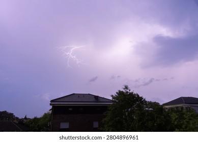 A stunning and dramatic lightning bolt illuminating a vibrant purple storm sky over a quiet suburban residential neighborhood during a summer evening thunderstorm.