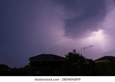 A stunning and dramatic night sky captured during a summer thunderstorm, featuring bright violet and purple lightning bolts splitting the dark clouds directly above suburban houses and trees.
