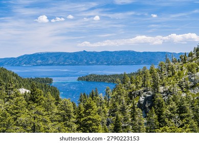 Stunning elevated panoramic view of Lake Tahoe, California, showcasing the iconic Emerald Bay with its brilliant turquoise and deep blue waters surrounded by lush green pine forests and granite rocky cliffs in the foreground. The vast alpine lake stretches to the horizon under a bright blue sky dotted with white cumulus clouds, with the Sierra Nevada mountain range visible in the background. This breathtaking summer landscape captures the pristine natural beauty of one of North America's most celebrated mountain lakes, ideal for travel, tourism, nature, and outdoor recreation content.
