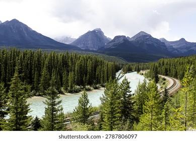 Stunning elevated view looking down over a historic railroad line winding through the dramatic mountain and river landscape of Banff National Park, Alberta, Canada. The railway tracks curve alongside a glacial turquoise river flanked by dense conifer forest, with the towering peaks of the Canadian Rockies rising majestically on all sides. This iconic transportation corridor through one of Canada's most celebrated national parks captures the remarkable contrast between human infrastructure and untamed wilderness. The sweeping bird's eye perspective emphasizes the sheer scale of the Rocky Mountain landscape dwarfing the railway below. A powerful image ideal for travel, transportation, Canadian wilderness, railroad heritage, adventure, and Rocky Mountain destination content.